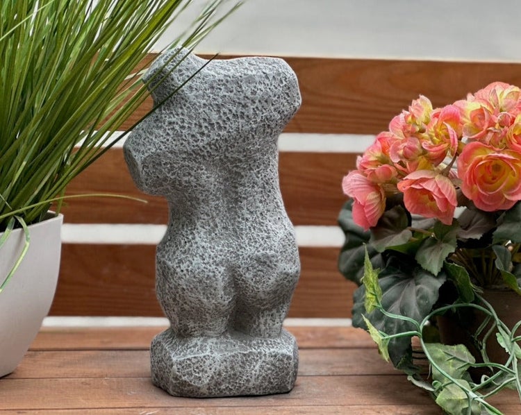 Dekorative Gartenfigur in Torso-Form aus grauem Kunststein mit strukturierter Oberfläche, stehend auf Holz zwischen Pflanzen.