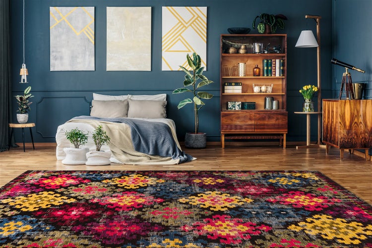 Bunter Teppich mit geometrischem Muster in einem Schlafzimmer mit dunkelblauer Wand, Holzmöbeln und Zimmerpflanzen.