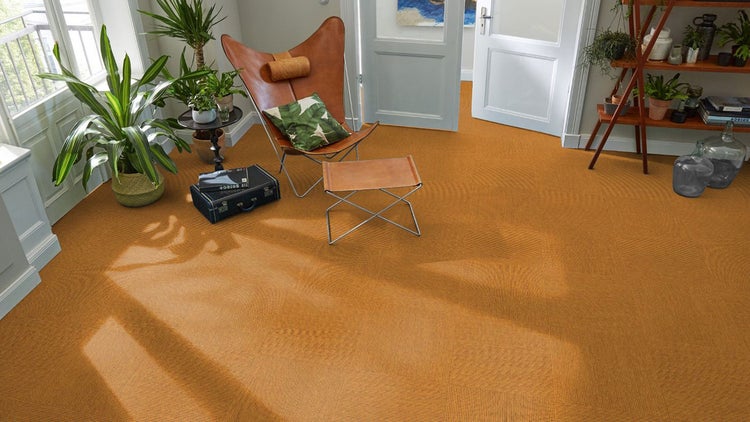 Wohnzimmer mit strukturierten Teppichfliesen im Schachbrettmuster, einem Butterfly-Sessel aus Leder, Pflanzen und hellem Lichteinfall.
