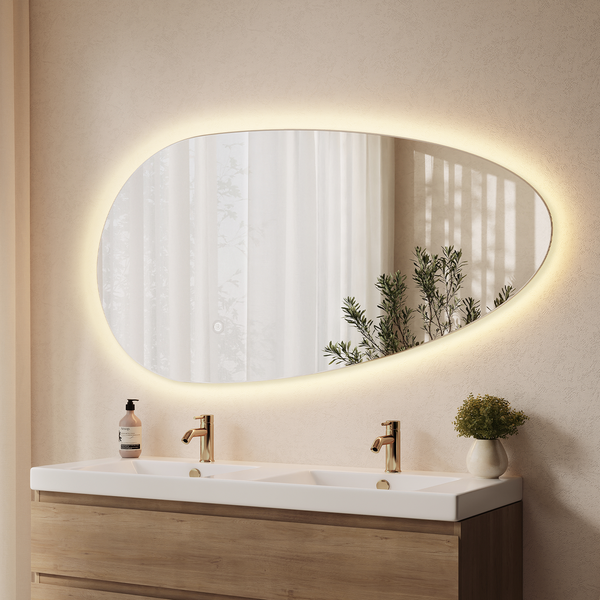 Organisch geformter Badezimmerspiegel mit indirekter LED-Beleuchtung über einem Waschtisch mit zwei goldenen Armaturen und Holzunterschrank.