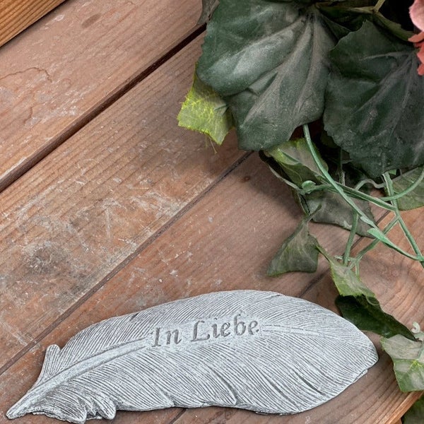 Grabschmuck in Form einer Feder aus Stein mit der Aufschrift In Liebe auf Holzdielen.