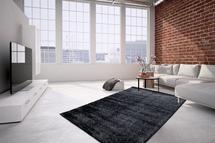 Großzügiges Wohnzimmer im Industrie-Stil mit dunklem Hochflor-Teppich, weißem Ecksofa, einer Ziegelwand und großen Fenstern.