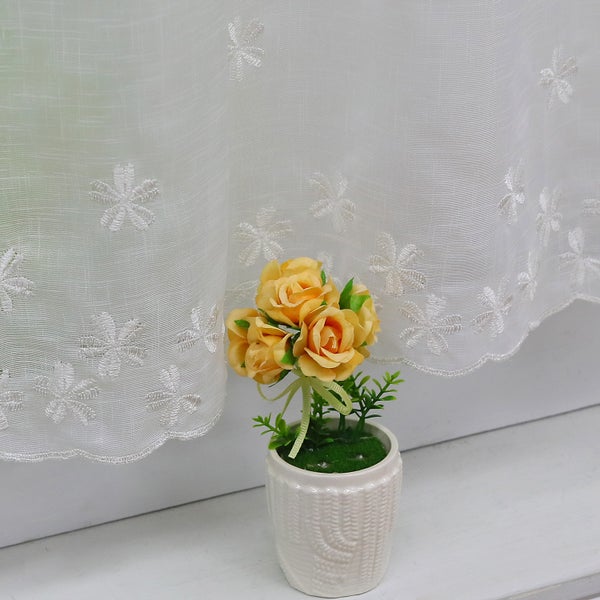 Dekorative Kunstblumen in einem Topf vor einem weißen Vorhang mit Blumenmuster.