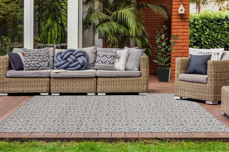 Moderner Außenteppich mit geometrischem Muster auf einer Holzterrasse vor einer Rattan-Lounge mit grauen Kissen.