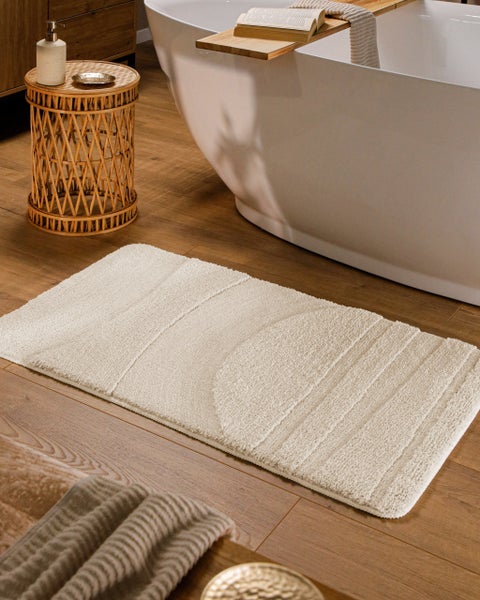Beige Badematte mit geometrischem Muster auf einem Holzfußboden vor einer weißen Badewanne in einem modern eingerichteten Badezimmer.