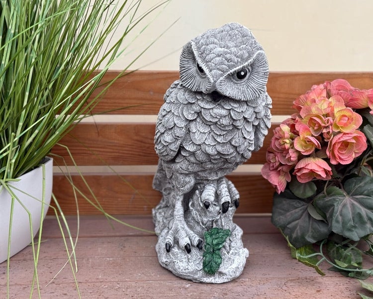 Gartenfigur Eule in Steinoptik auf einer Holzbank, umgeben von Pflanzen.