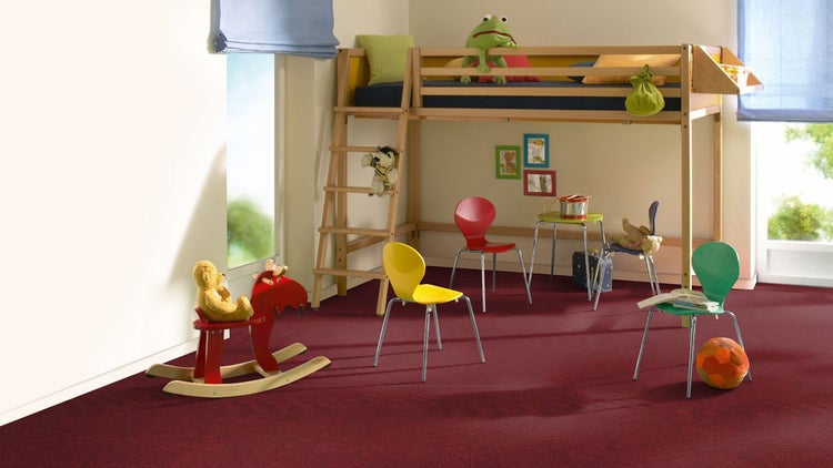 Helles Kinderzimmer mit rotem Teppichboden, einem Hochbett aus Holz, bunten Stühlen und einem Schaukelpferd.