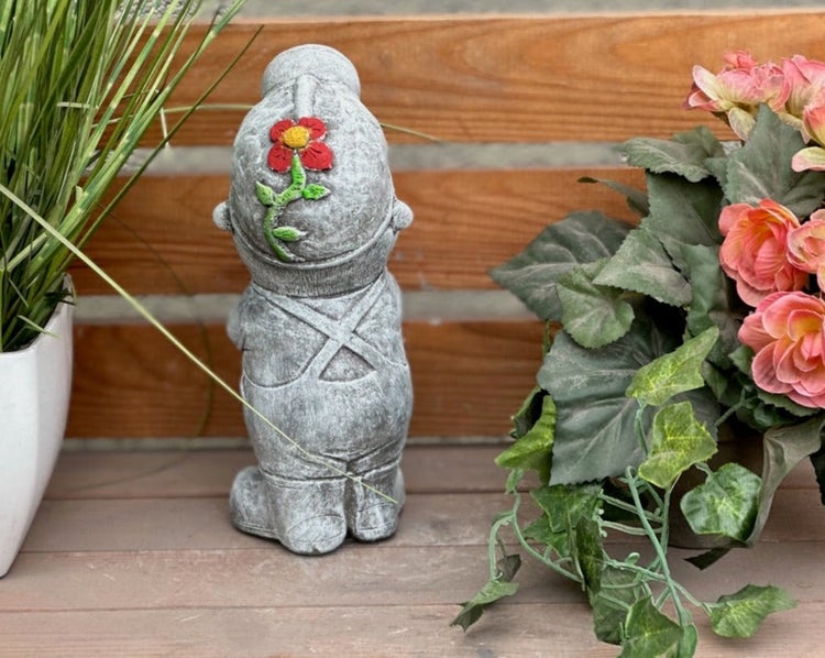 Gartenzwerg-Figur aus Stein in der Rückansicht mit einer Blume auf der Mütze, platziert zwischen Pflanzen.