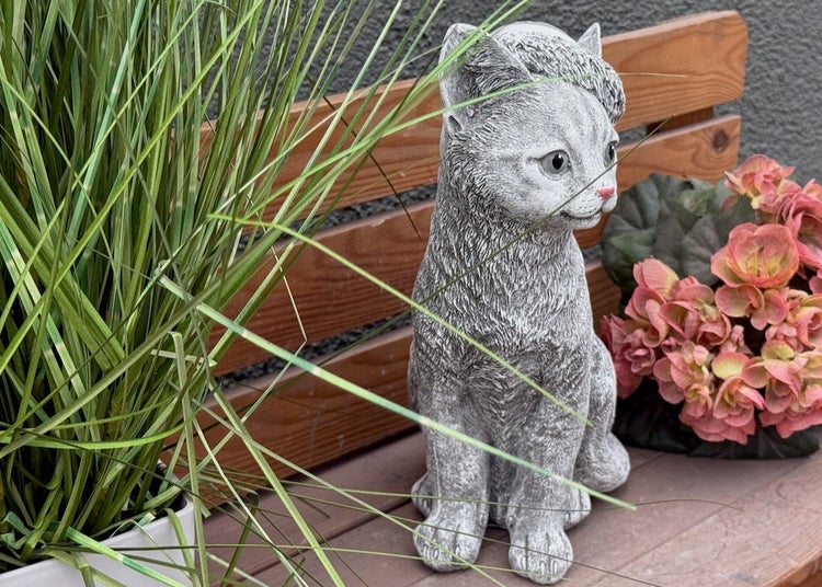 Dekorative Katzenfigur aus Stein für den Garten auf einer Holzbank neben Gräsern und Blumen.