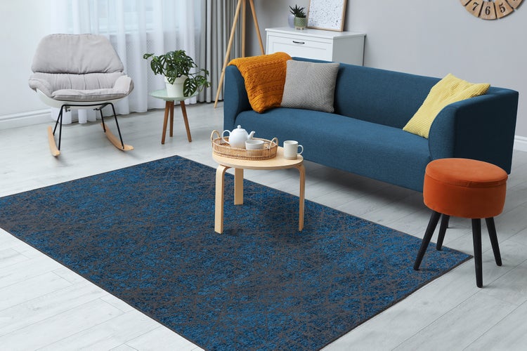 Hell eingerichtetes Wohnzimmer mit einem blau-grau gemusterten Teppich, einem blauen Sofa und einem hellen Holztisch.