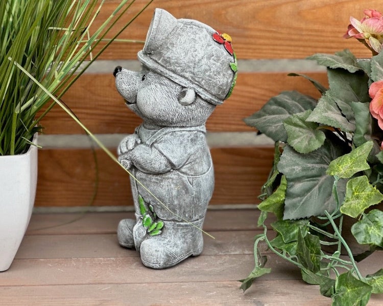 Graue Gartenfigur aus Steinguss in Form eines niedlichen Bergmanns mit Helm und Overalls, stehend auf Holzfläche zwischen Pflanzen.
