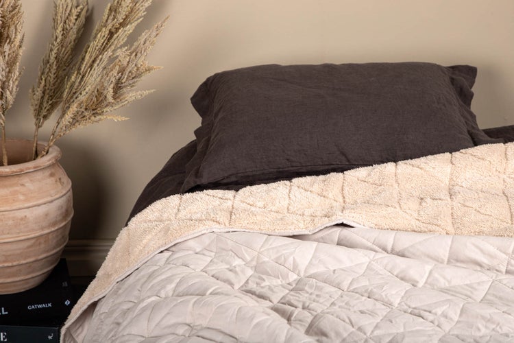 Bett mit dunkelbraunem Kissen und hellbeiger Steppdecke. Daneben steht eine große Tonvase mit getrocknetem Ziergras.