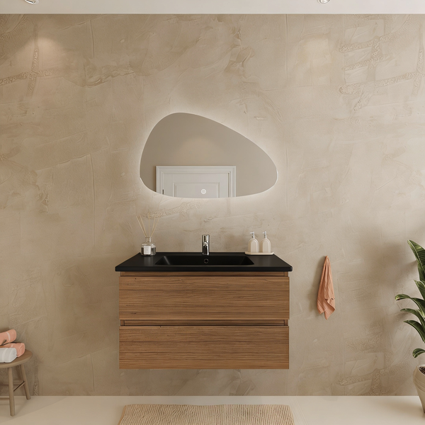 Moderner Waschplatz mit holzfarbenem Unterschrank, schwarzem Waschtisch und asymmetrischem beleuchtetem Spiegel vor beige strukturierter Wand.