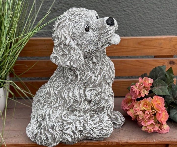 Gartenfigur in Form eines sitzenden Hundes aus Stein mit lockigem Fell auf einer Holzbank neben Blumen.