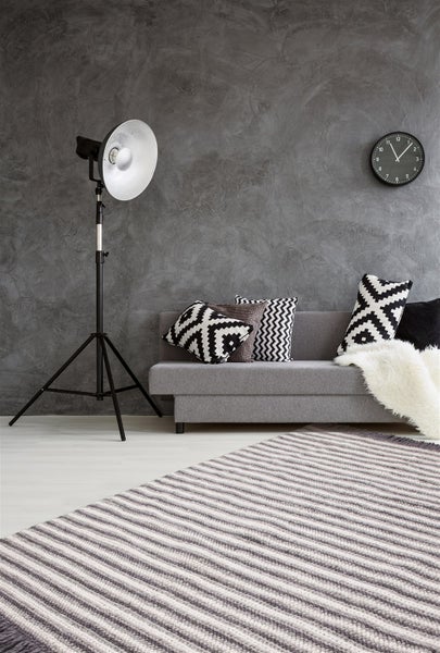 Modernes Wohnzimmer mit grauer Strukturwand, grauem Sofa mit Dekokissen, Studioleuchte auf Stativ, Wanduhr und gestreiftem Teppich.