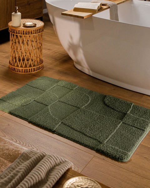 Grüner Badevorleger mit geometrischem Karomuster auf Holzboden neben einer freistehenden Badewanne und einem geflochtenen Beistelltisch.