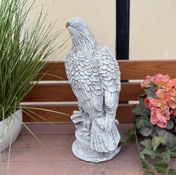 Gartenfigur Adler in Steinoptik auf einer Holzbank, umgeben von Zierpflanzen.