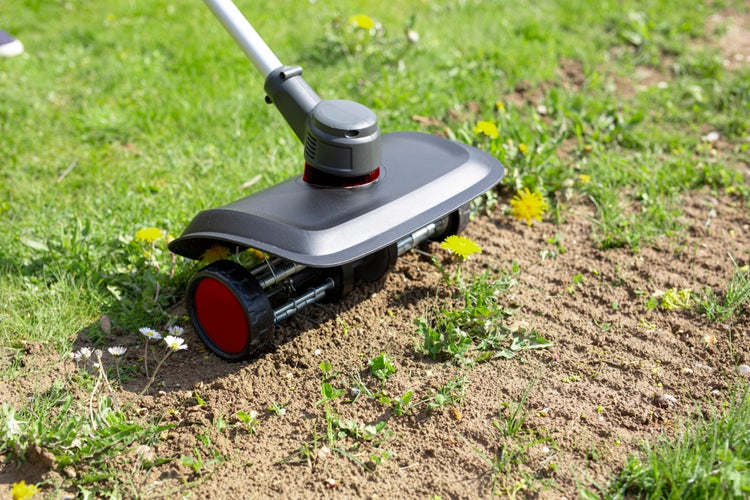 Elektrischer Motorhacken-Aufsatz im Einsatz auf Gartenboden zur Unkrautentfernung zwischen Gras und Wildblumen.