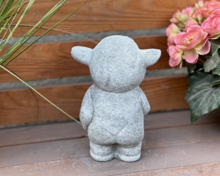 Rückansicht einer kleinen stehenden Gartenfigur in Lammform aus Stein auf Holzuntergrund.