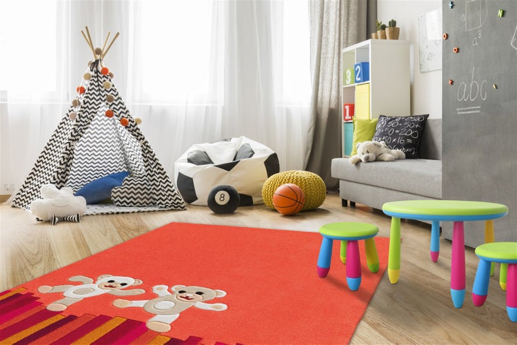 Heller Kinderzimmer-Teppich in Rot mit Teddybär-Motiven und bunten Streifen, umgeben von Spielzelt, Sitzsack und bunten Kindermöbeln.
