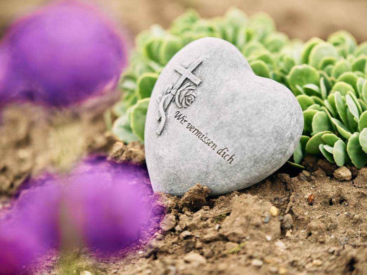Grabschmuck in Herzform aus Stein mit eingraviertem Kreuz, Rose und Inschrift Wir vermissen dich, platziert auf einem Grab.