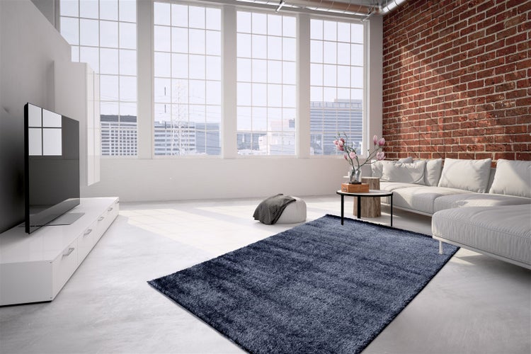 Helles, geräumiges Wohnzimmer im Industriestil mit einem blauen Hochflor-Teppich, weißem Sofa, einer Ziegelwand und großen Fenstern.