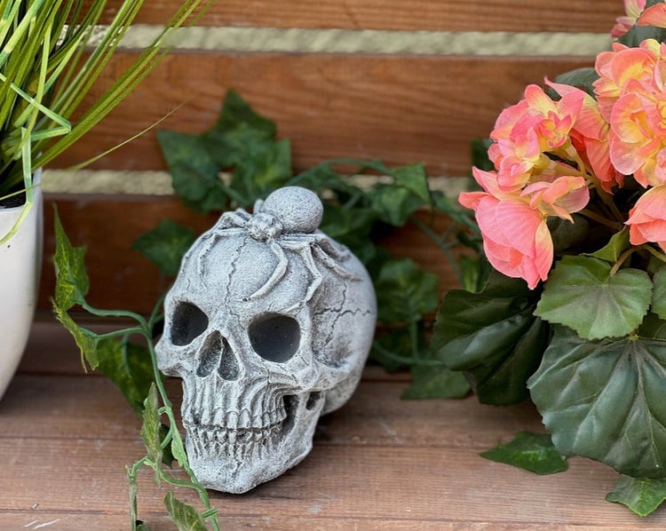 Dekomotiv Totenkopf mit Spinne aus Steinguss, platziert auf Holz zwischen grünen Pflanzen und Blüten.