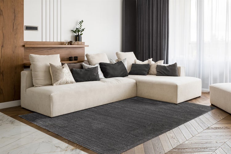 Großes Wohnzimmer mit hellem Ecksofa, dunkelgrauem Teppich auf Fischgrät-Parkett, Holzregal und Vorhängen. Hell und modern eingerichtet.