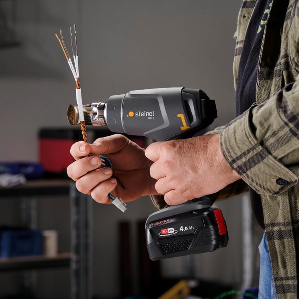 Person verwendet ein akkubetriebenes Steinel Heißluftgebläse MH 1 mit einem Bosch 18 Volt Akku zur Bearbeitung von isolierten elektrischen Kabeln.
