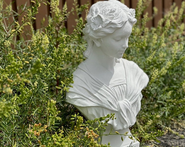 Weiße Gartenbüste einer Frau mit Blumen im Haar, umgeben von grünen Pflanzen im Garten.