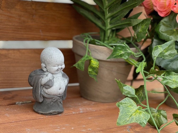 Kleine Buddha Gartenfigur aus Stein auf einer Holzoberfläche neben einem Blumentopf mit Pflanze als Dekoration.