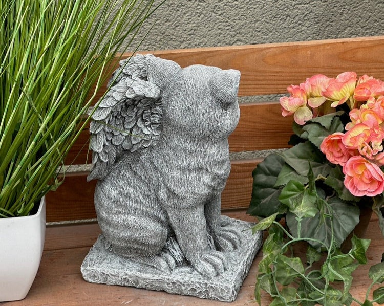 Gartenfigur Katze mit Engelsflügeln aus Steinguss, sitzend auf einer Holzbank neben Pflanzen.