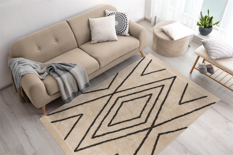 Hell gestaltetes Wohnzimmer mit einem rechteckigen Teppich mit geometrischem Rautenmuster und Fransen auf hellem Boden neben einem Sofa.