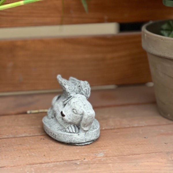 Kleine Gartenfigur aus Stein in Form eines liegenden Hundes mit Engelsflügeln als Dekoration für den Außenbereich.