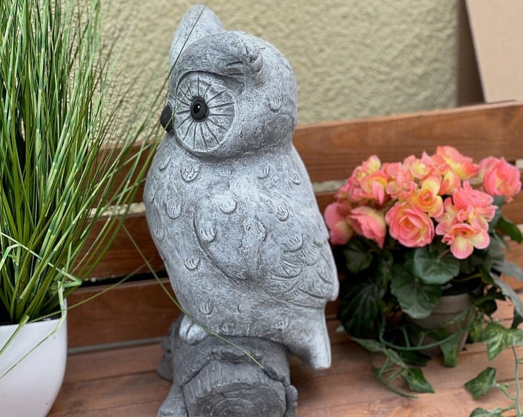 Gartenfigur Eule aus Steinguss auf einem Ast sitzend, dekoriert auf einer Holzbank neben Pflanzen.