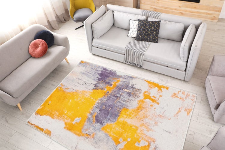 Modernes Wohnzimmer mit grauem Sofa, gelbem Sessel und einem rechteckigen Teppich mit abstraktem gelb-violettem Muster auf Holzboden.