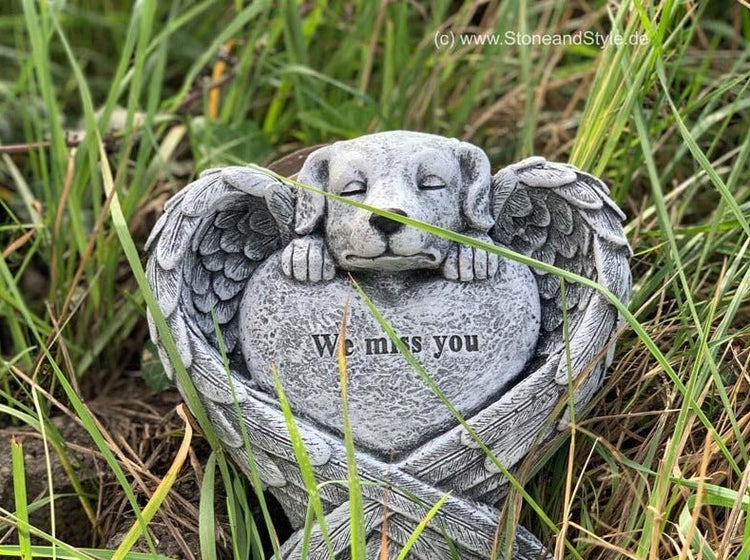Grabschmuck in Form eines Hundes mit Engelsflügeln, der auf einem Herz mit der Inschrift We miss you liegt, aus witterungsbeständigem Steinguss.