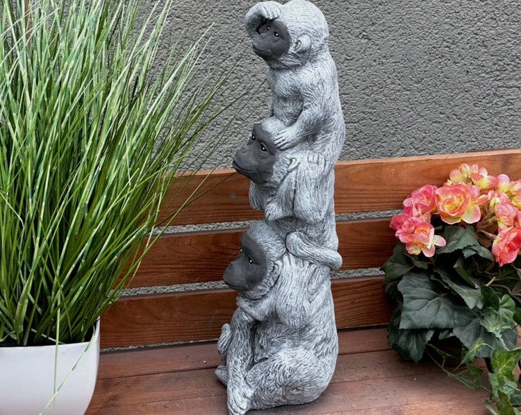 Gartenfigur drei weise Affen übereinander aus Steinguss, auf einer Holzbank neben Topfpflanzen dekoriert.