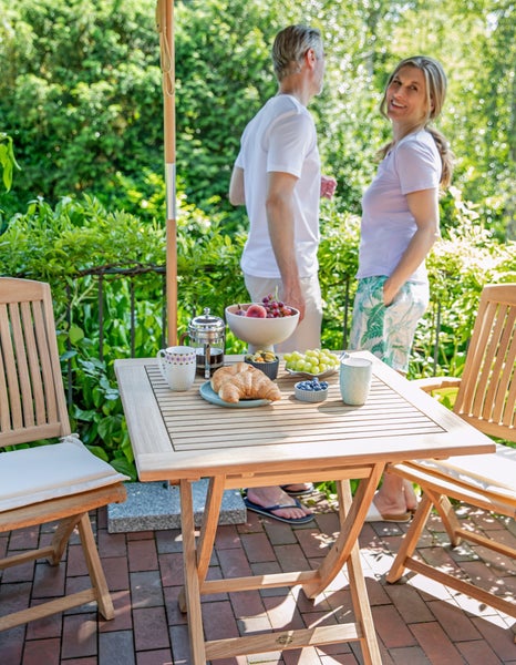Gartenszene mit Tisch, Stühlen, Sonnenschirm und Personen im Hintergrund.