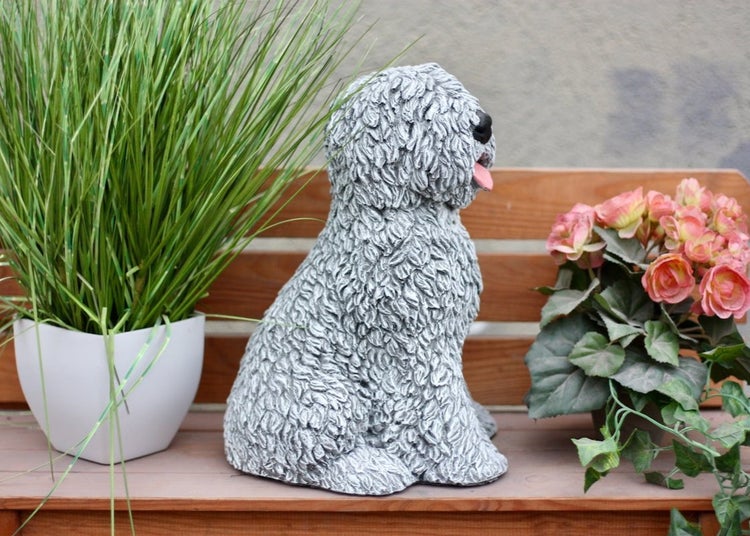 Gartenfigur Hund mit lockigem Fell sitzend auf einer Holzbank zwischen Topfpflanzen.