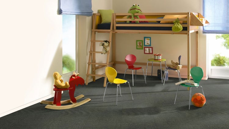 Dunkelgraue Teppichfliesen in einem hellen Kinderzimmer mit Hochbett aus Holz, bunten Stühlen und Spielzeug.