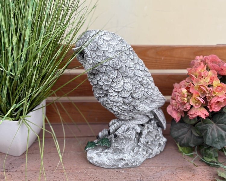 Gartenfigur in Form einer detailreichen Eule aus Stein, platziert auf einer Holzbank zwischen einer Pflanze und Blumen.