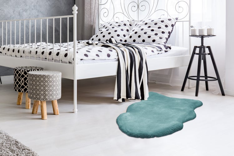 Türkisfarbener Kunstfellteppich in Lammfellform vor einem weißen Metallbett in einem hellen Schlafzimmer.
