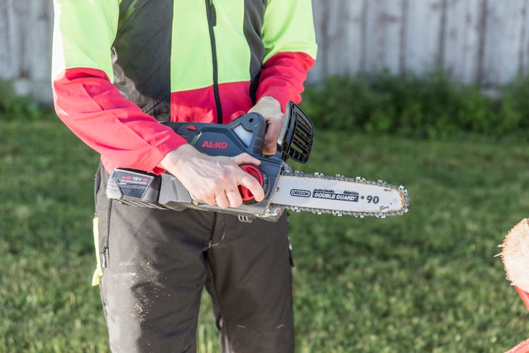 Eine Person hält eine AL-KO Akku-Kettensäge im Garten, ausgestattet mit einem Oregon-Schwert und einem AL-KO 18-Volt-Akku.