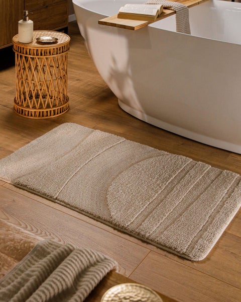 Beige Badematte mit Reliefmuster auf Holzfußboden neben freistehender Badewanne und geflochtenem Beistelltisch im hellen Badezimmer.