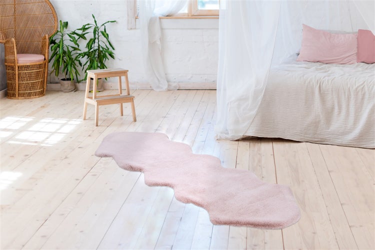 Rosa Kunstfell-Läufer in unregelmäßiger Form auf hellem Holzboden in einem hellen Schlafzimmer mit Bett und Korbsessel.