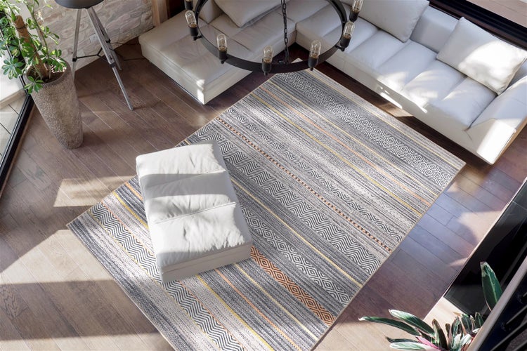 Moderner Wohnbereich mit großem gemustertem Teppich in Grau und Gelb auf dunklem Holzboden. Weißes Ledersofa, Hocker und helle Atmosphäre.