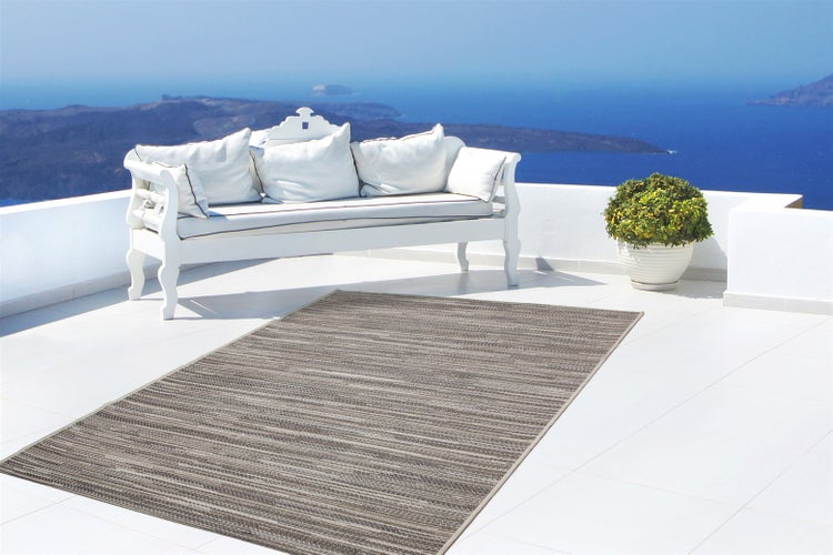 Outdoor-Teppich mit Streifenmuster auf einer hellen Terrasse mit weißer Sitzbank, Pflanzgefäß und Meerblick im Hintergrund.
