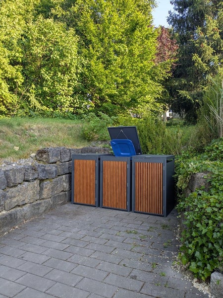 Moderne 3er Mülltonnenbox aus dunklem Metall und Holzlatten auf gepflastertem Boden im Garten. Ein Deckel ist geöffnet.