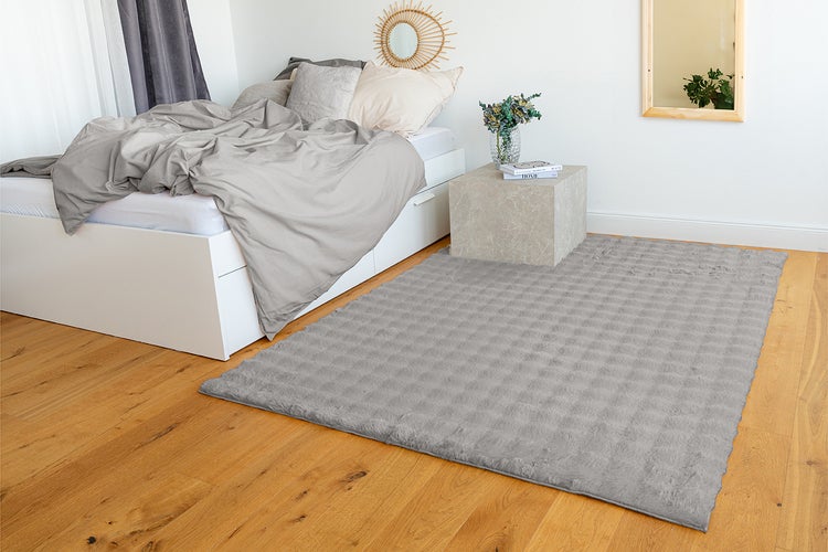 Rechteckiger, grauer Teppich mit Waffelstruktur in einem hellen Schlafzimmer auf Parkettboden neben einem weißen Bett.